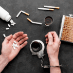 Vue de dessus de deux mains d’une personne menottée, entourée de cigarettes, de comprimés, de verres d’alcool et d’une flasque. Scène illustrant les addictions.