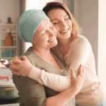 Une femme souriante atteinte d'un cancer portant un foulard médical est tendrement enlacée par une autre femme souriante.
